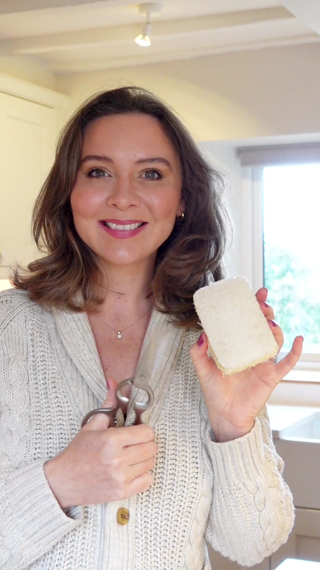Woman with brown hair smiling and holding up Seep Sponge Scourer in left hand and scissors in the right hand