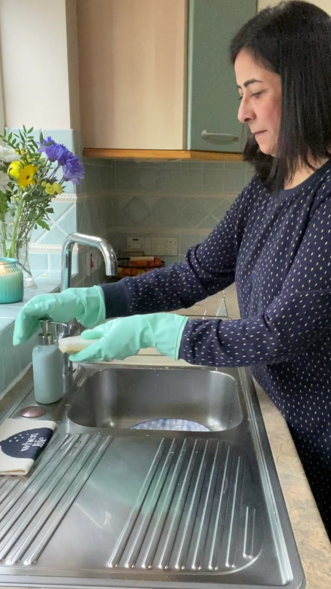 Woman with short black hair at kitchen sink wearing Seep Eco Rubber Gloves pumping washing up liquid onto a white Seep Sponge Scourer