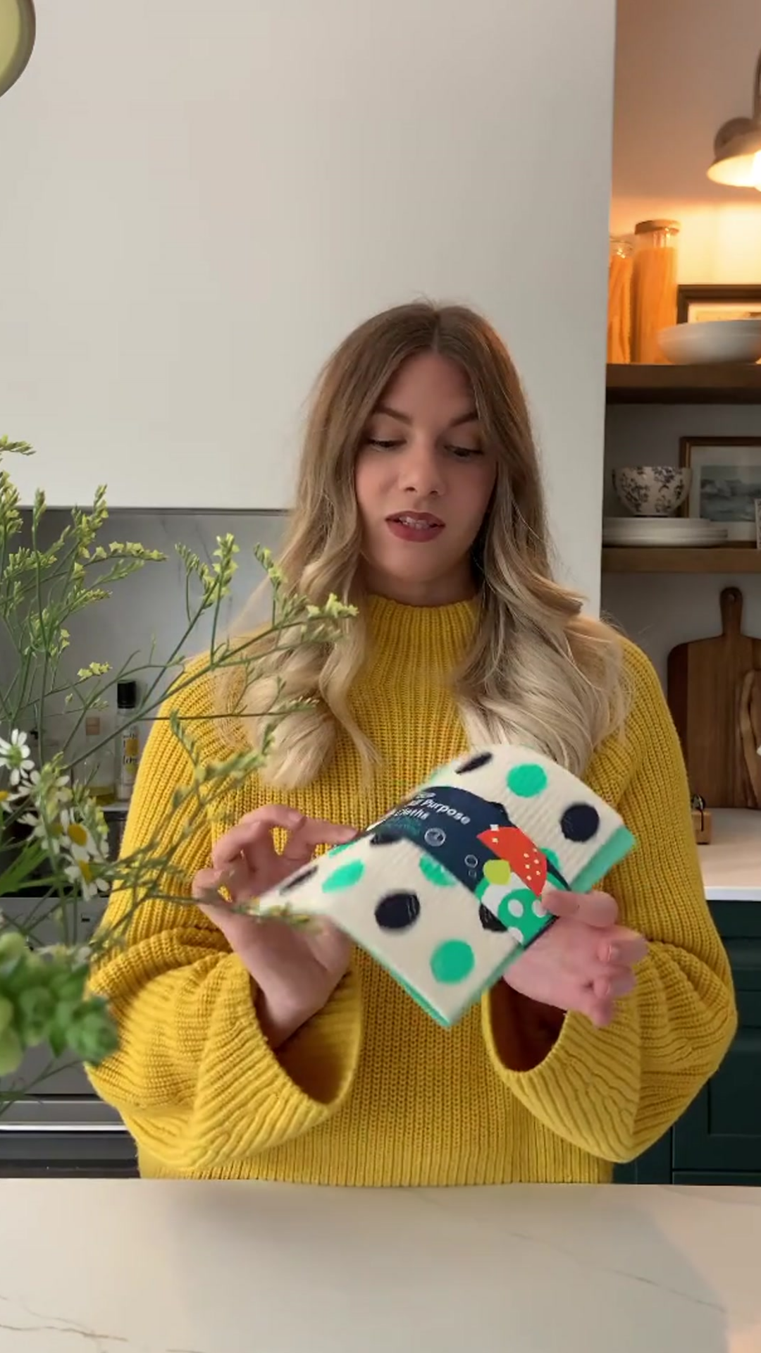 Woman with dark blond hair and yellow sweater standing behind a kitchen counter holding up a pack of Seep Eco Sponge Cloths