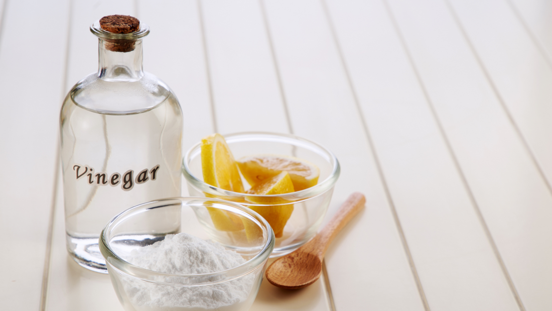Distilled vinegar in a glass bottle with lemon and baking soda in small bowls next to vinegar bottle